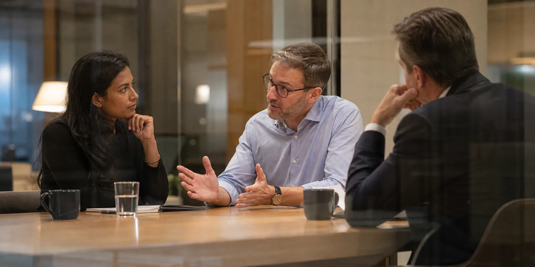 Three audit and quality colleagues rehearse a difficult client conversation around a wooden table in a warm glass-walled office.