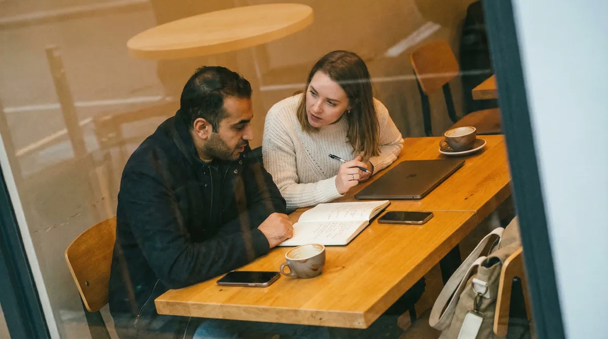 Two colleagues in a quiet modern office, one leaning toward the other across a wooden table during an informal conversation, natural window light, candid film photography style