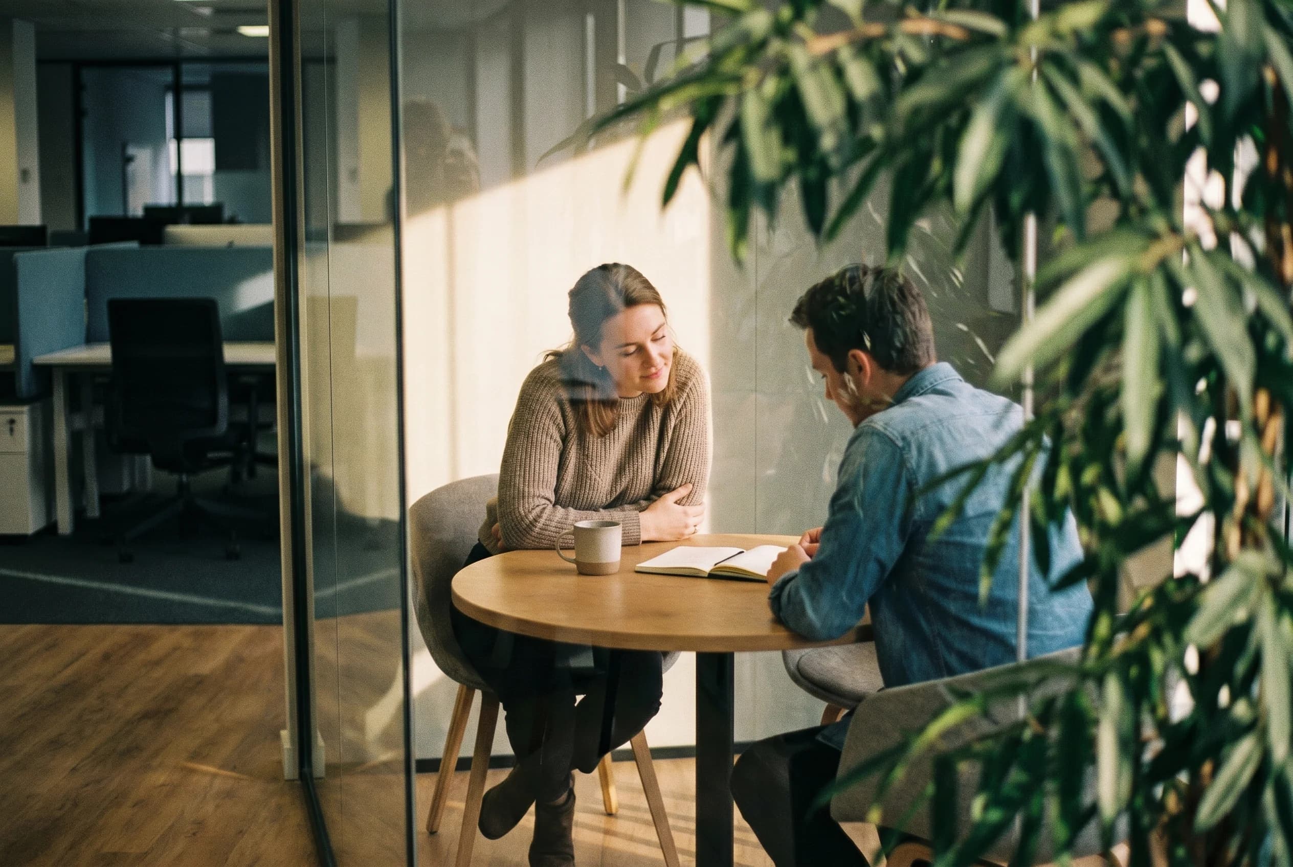 Two account team colleagues rehearse a difficult customer conversation at a small table in a warm glass-walled office, photographed candidly through a plant.
